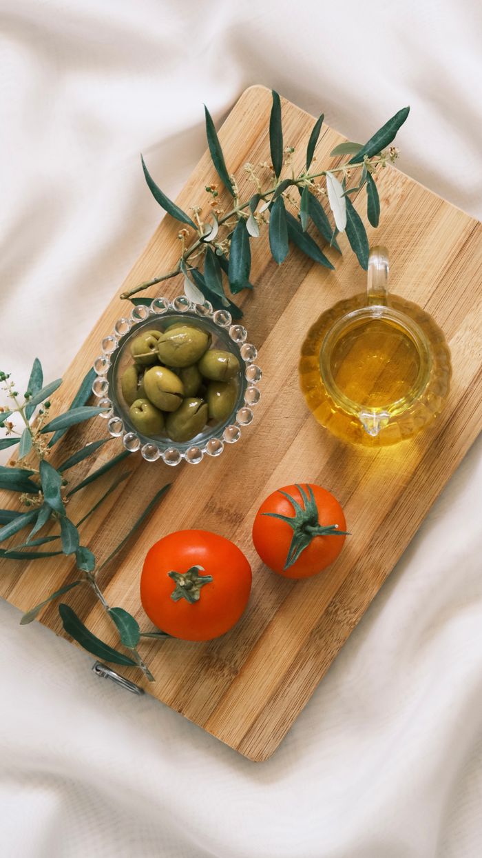 hero-img-02 Overhead view of fresh tomatoes, olives, and olive oil on a wooden cutting board surrounded by olive branches.